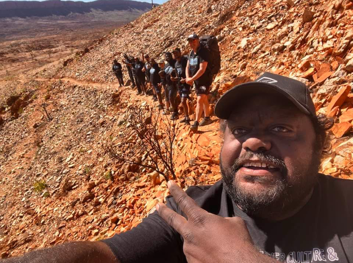 Benjamin Kenny, Western Arrernte man and 100% Finke River Trip Leader, captures a joyful moment with travellers on the rugged Larapinta Trail. Walking on Country, sharing stories, and connecting with the ancient landscapes of Tjoritja (West MacDonnell Ranges) with Inspiration Outdoors.