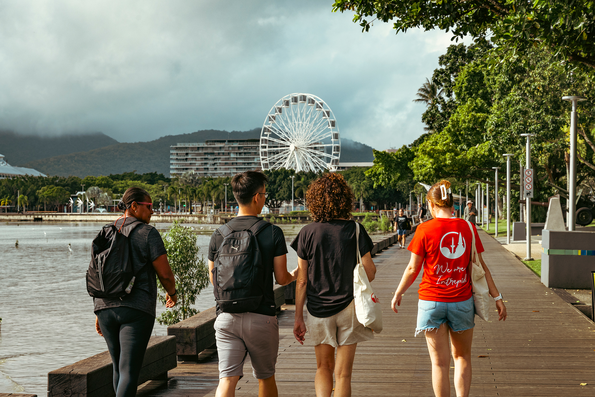 A small group of travellers from Inspiration Outdoors stroll along the scenic Cairns boardwalk in Queensland, Australia, with the iconic Ferris wheel and lush mountains in the background.