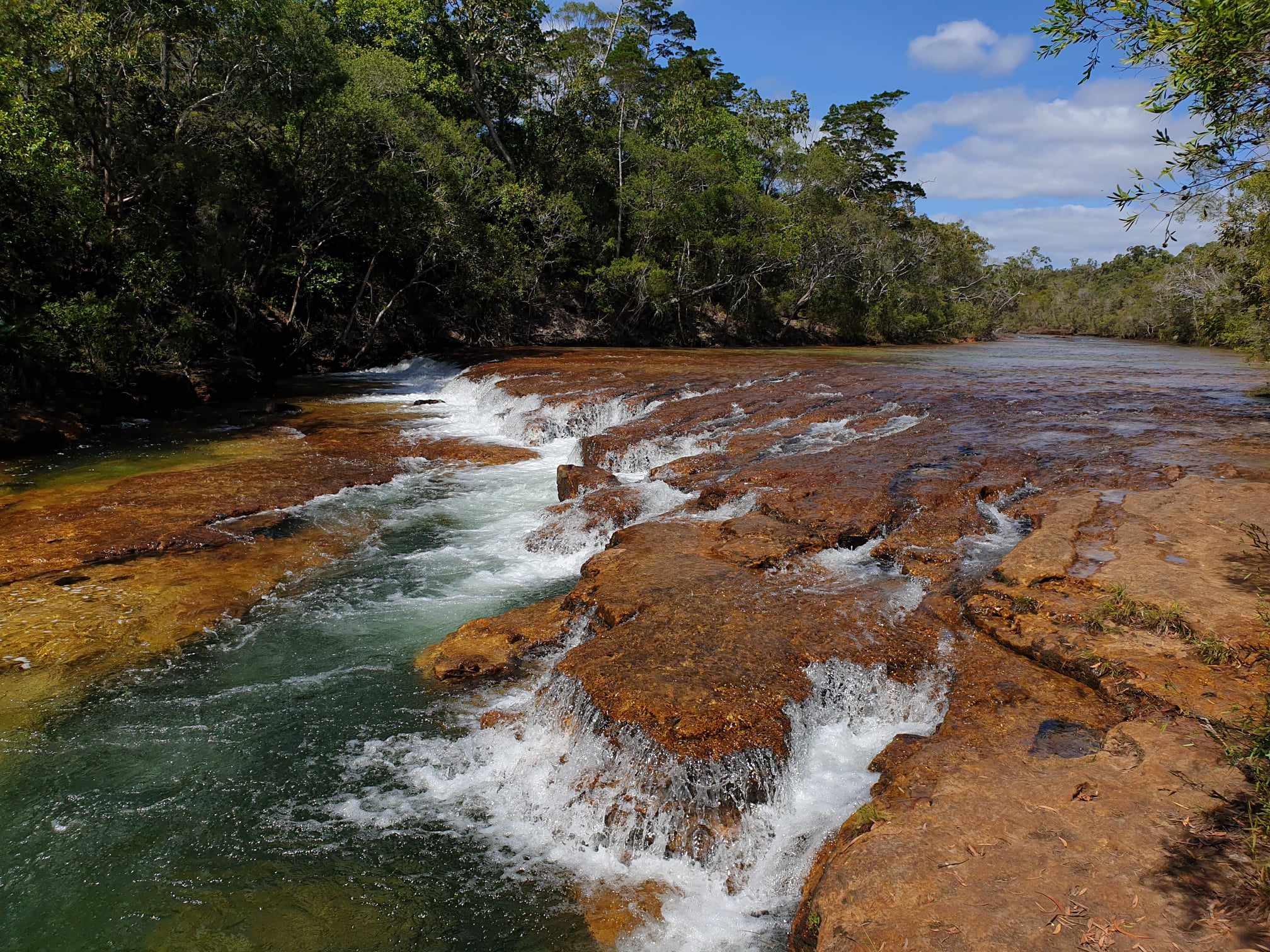 A stunning view of Eliot River in Cape York, Queensland, showcasing clear water cascading over natural rock formations surrounded by lush green forest under a bright blue sky with inspiration outdoors