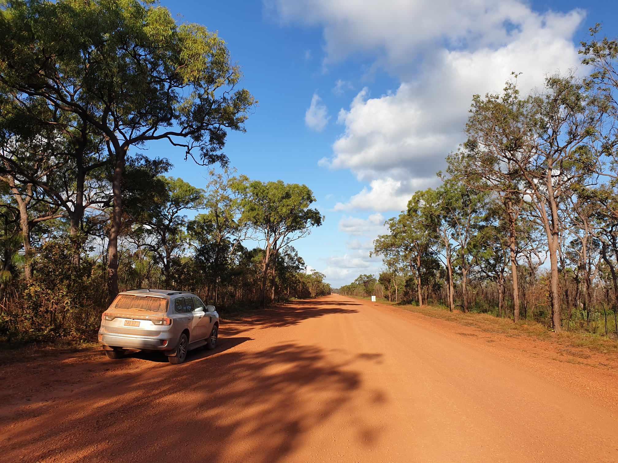 A dusty 4WD vehicle parked along a red dirt road near Jardine River in Cape York, Queensland, surrounded by tall trees under a bright blue sky with scattered clouds with inspiration outdoors