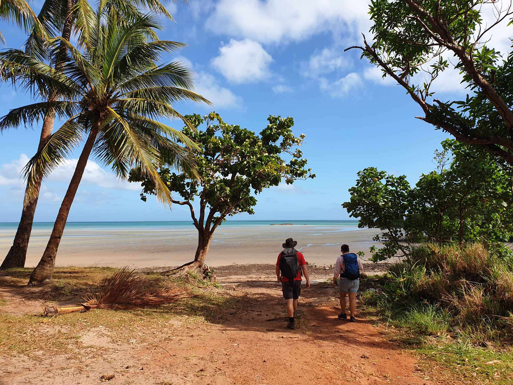 Two hikers with backpacks walking towards the serene, sandy beach at Pajinka, Cape York, Queensland, surrounded by palm trees and lush greenery under a bright blue sky with inspiration outdoors